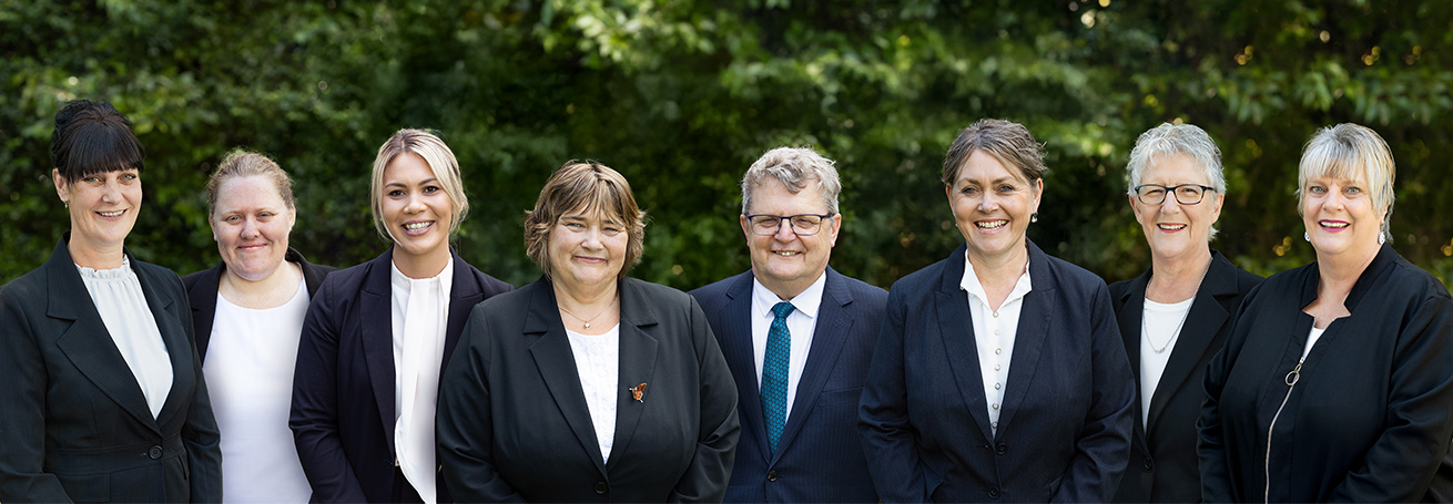 The team of eight staff members from a funeral services company standing outdoors in formal attire, smiling at the camera with greenery behind them.
